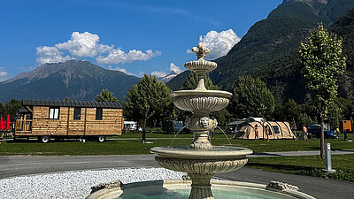 Steinbrunnen im Vordergrund, dahinter Holzmobilheim, Zelte und Berge unter blauem Himmel auf einem Campingplatz